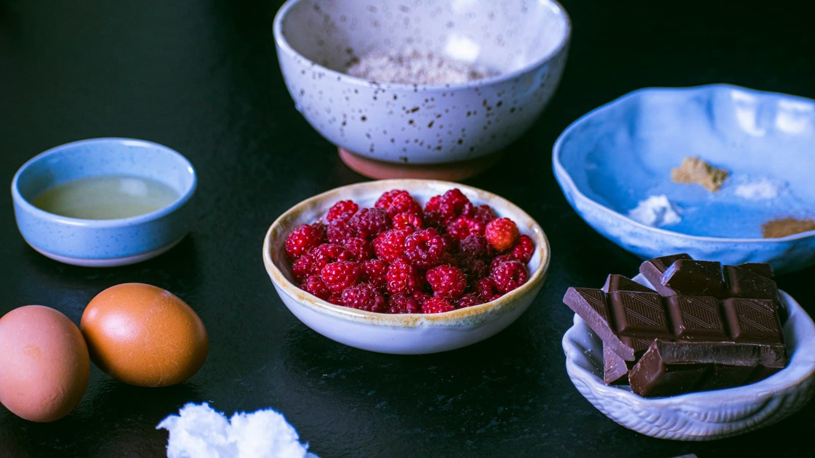 Baking ingredients on a dark counter: two brown eggs, a bowl of raspberries, pieces of dark chocolate and bowls of flour and spices