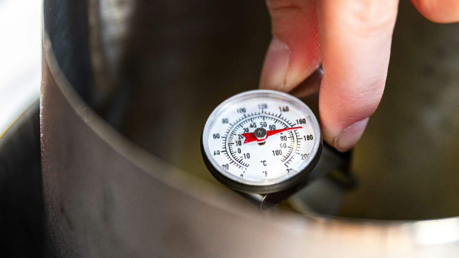 A hand holding a dial thermometer in a steel pot, measuring temperature in a kitchen