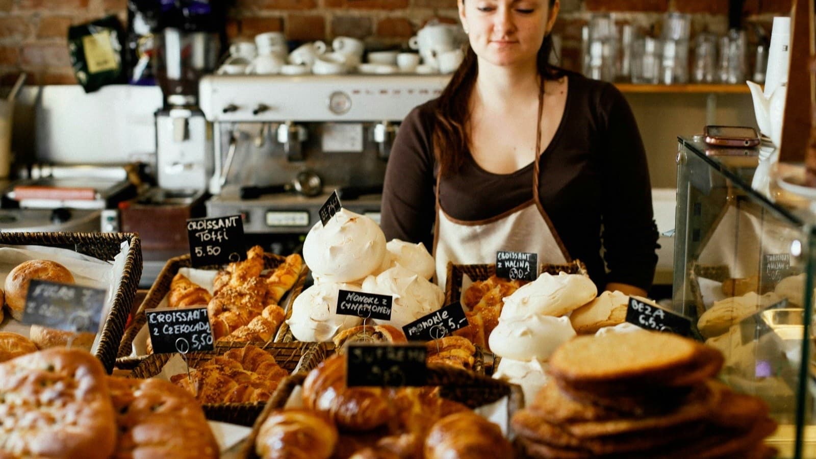 An owner in an apron behind a pastry shop counter full of croissants, meringues and sweet buns with price tags