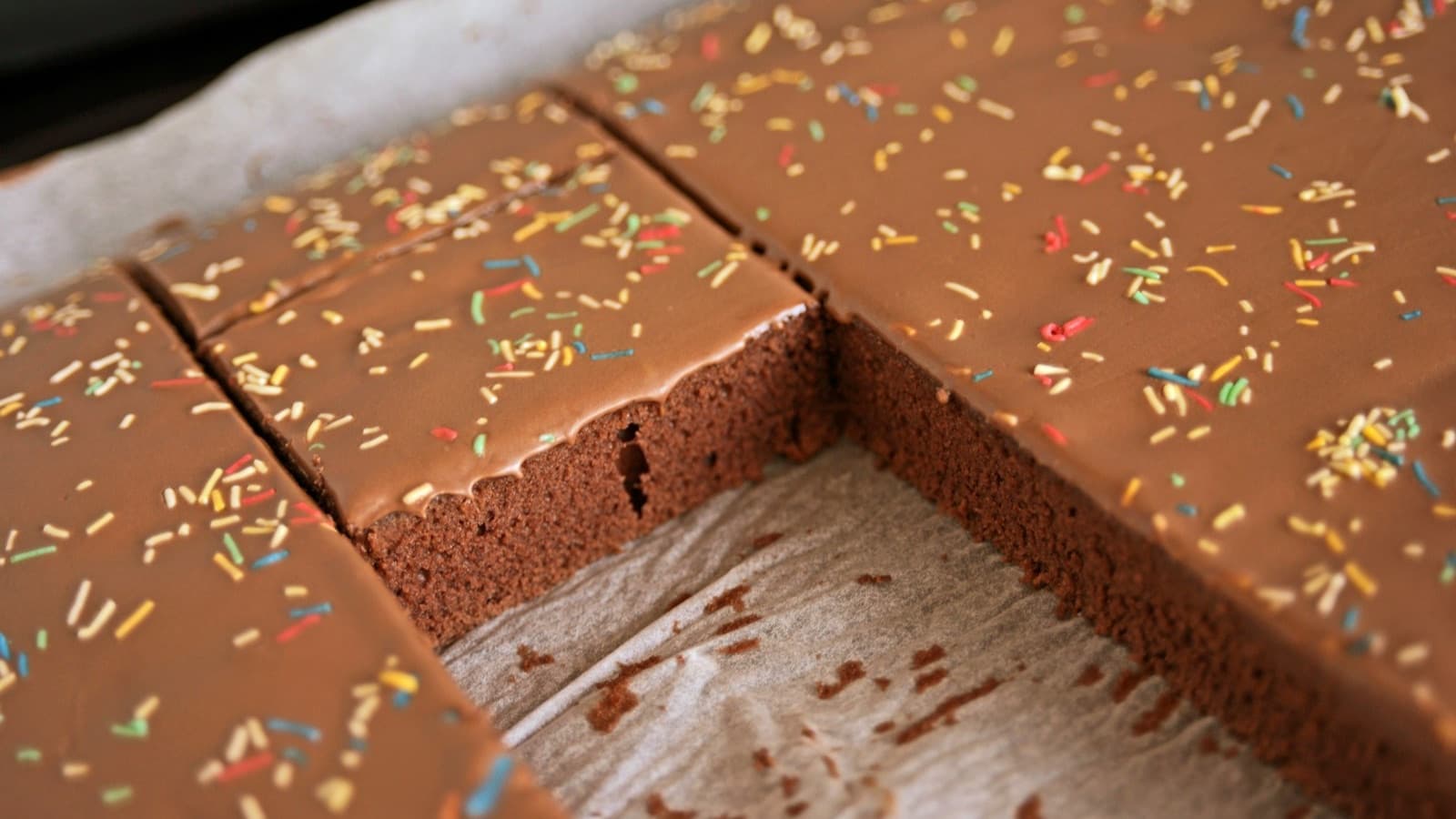 A chocolate cake with sprinkles on a tray lined with baking paper, cut into square slices