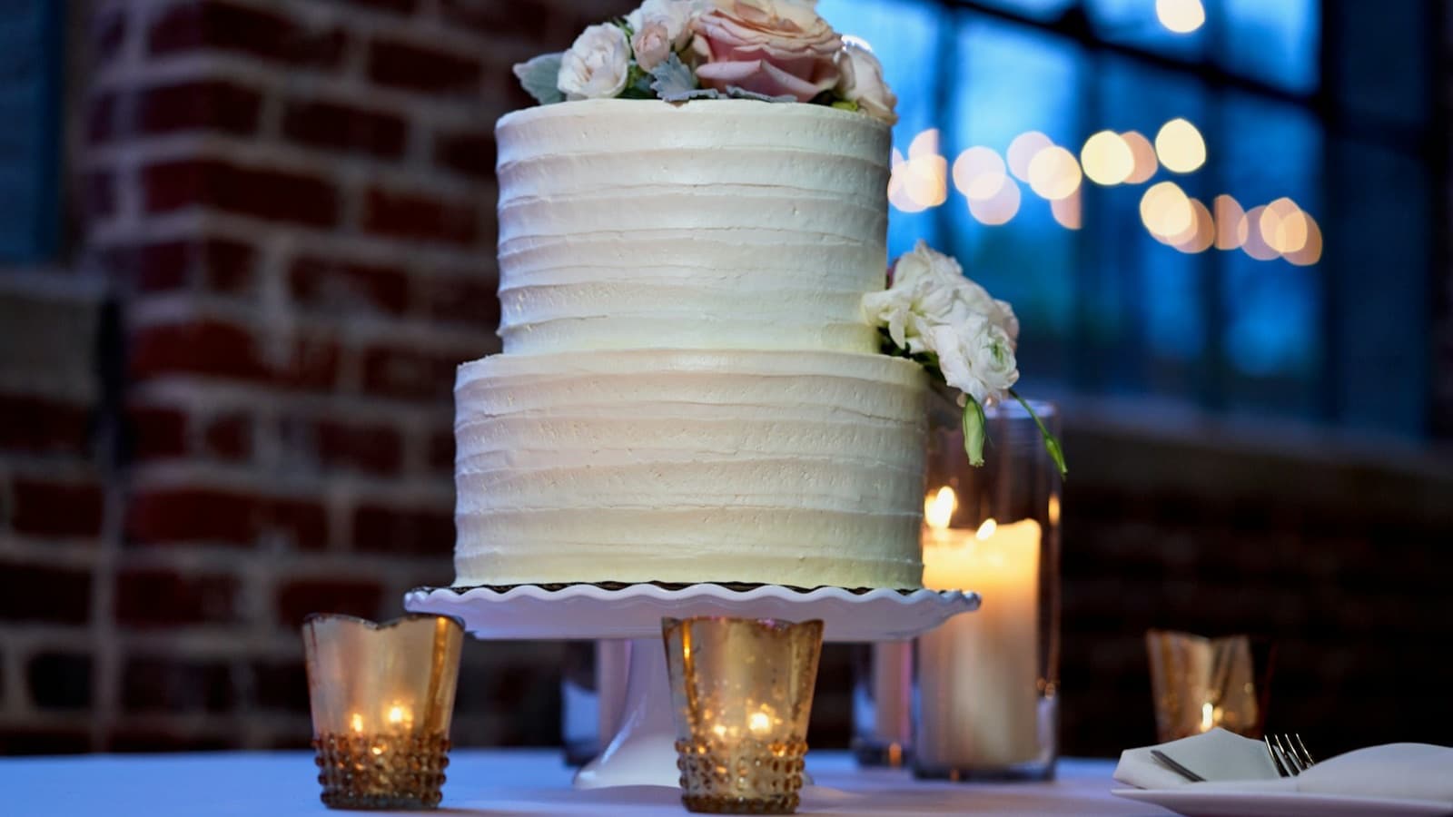 A white two-tier cake with roses on a cake stand, surrounded by candles, a celebration cake for a party