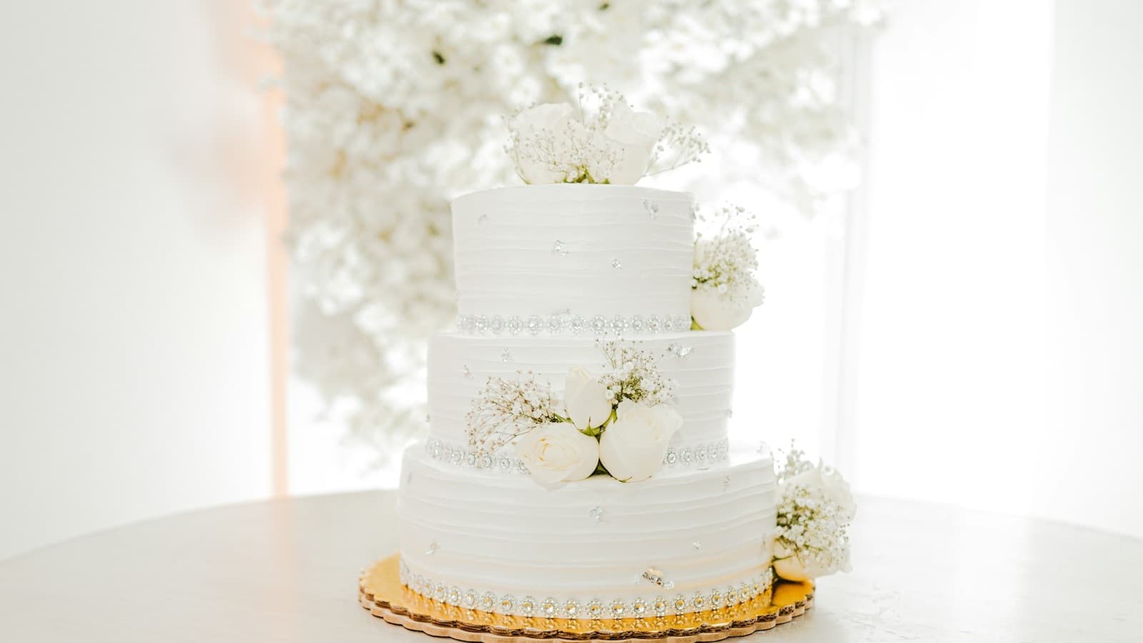 A three-tier white wedding cake with white roses and baby's breath on a gold cake board